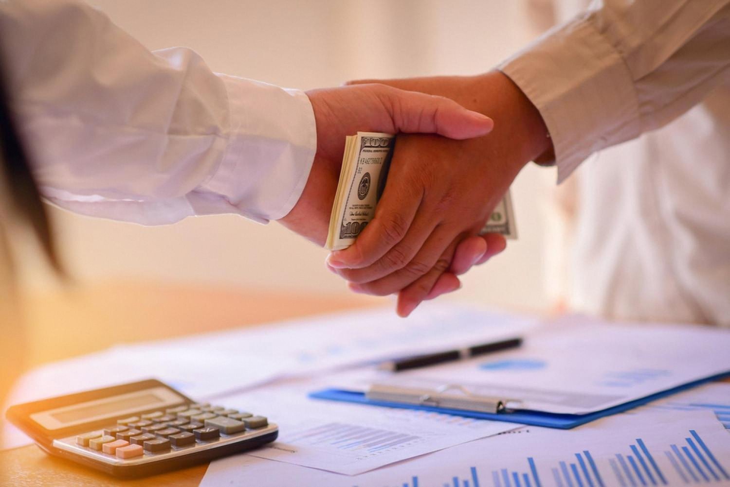Close-up of two hands exchanging cash over financial reports and a calculator, illustrating a kickback scheme inside a company